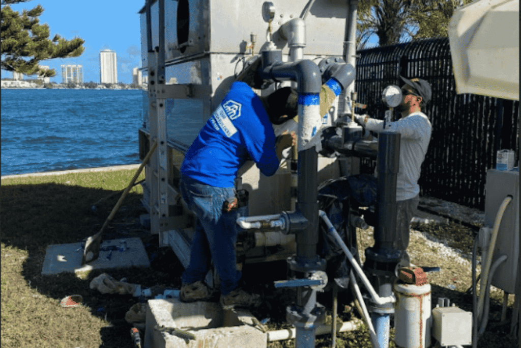 Commercial Plumbing System Maintenance and Repair Two licensed plumbers performing maintenance on an outdoor commercial plumbing and pump system near a waterfront property.