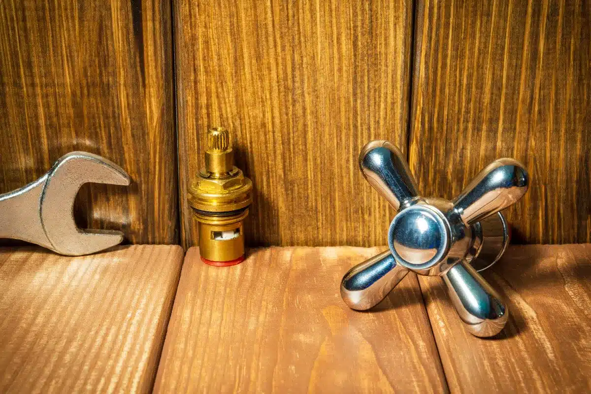 Metal faucet handle, brass valve cartridge, and adjustable wrench placed on a wooden surface, representing plumbing repair and faucet part replacement.