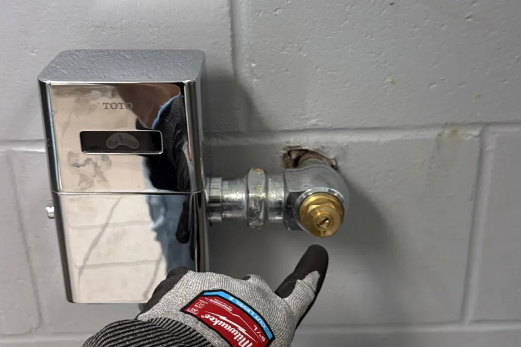 Plumber inspecting and adjusting a commercial toilet flush valve and shutoff connection on restroom wall.