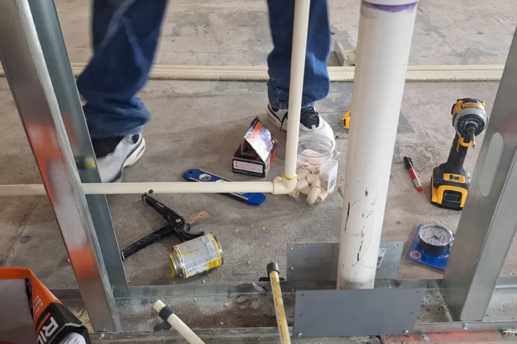 PVC plumbing rough-in installation inside framed wall with tools and fittings on construction site floor.