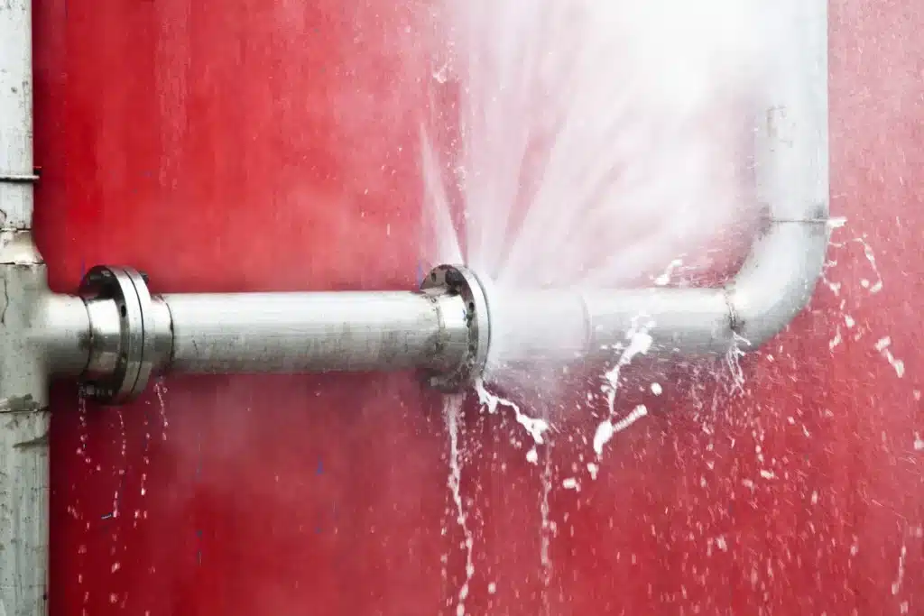 Metal plumbing pipe with a burst joint spraying water against a wall, showing severe leak damage requiring immediate repair.