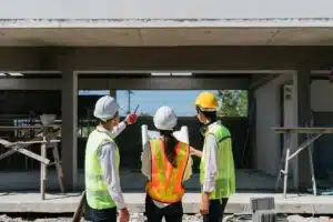 Construction Team Reviewing Plans at Residential Job Site Three construction workers wearing hard hats and safety vests reviewing blueprints and discussing progress in front of a residential building under construction.