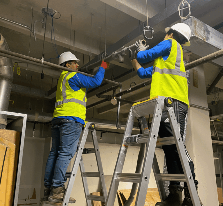 Two licensed plumbing contractors wearing white hard hats and high-visibility yellow vests stand on aluminum A-frame ladders to install and secure a large grey pipe into an exposed industrial ceiling.