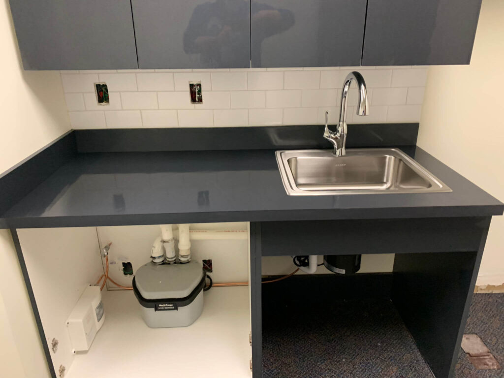 Commercial breakroom sink with stainless steel basin, dark countertop, subway tile backsplash, and visible plumbing and pump system under cabinet.